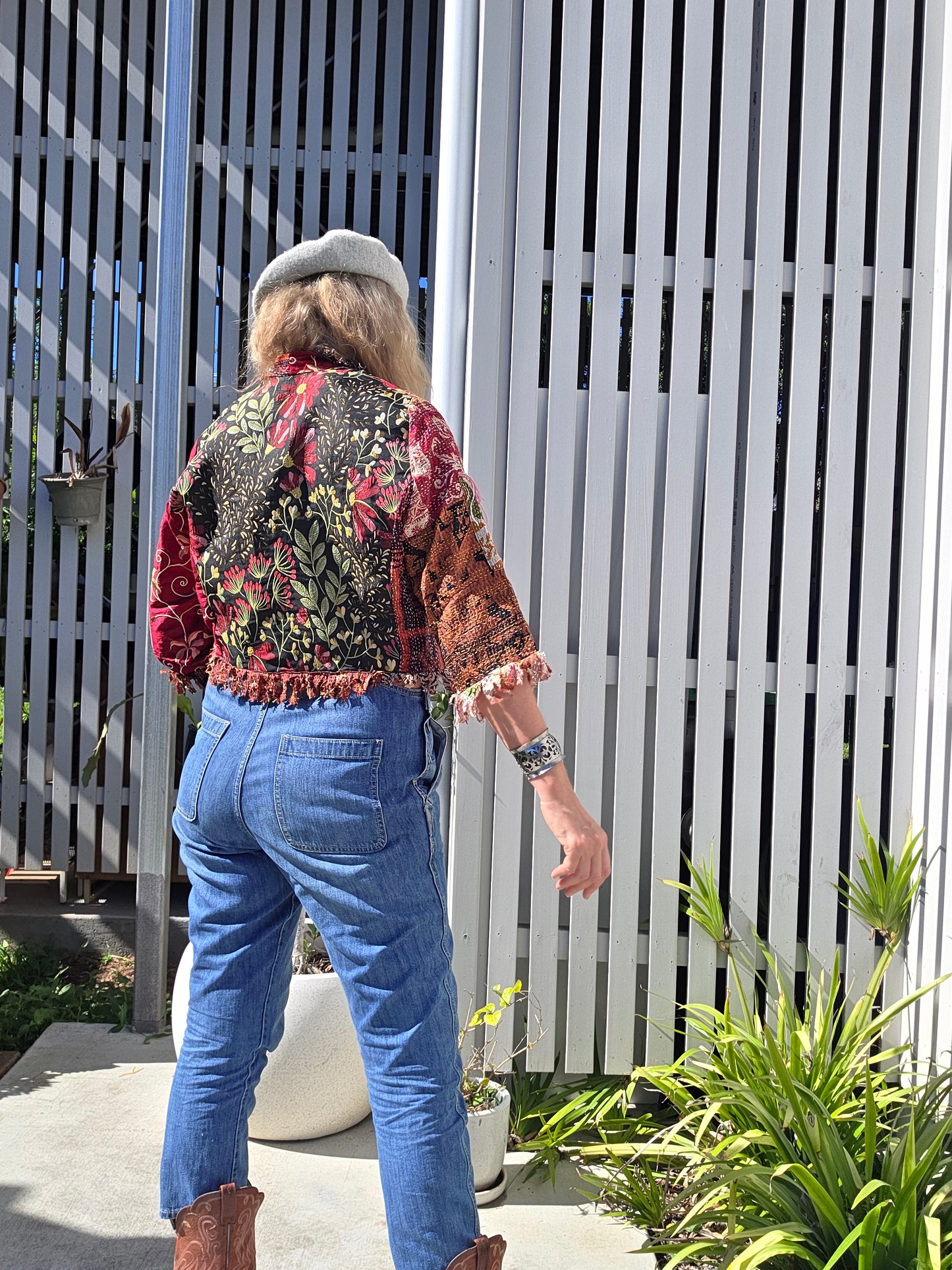 Person wearing a colorful patterned jacket and blue jeans standing against a white fence