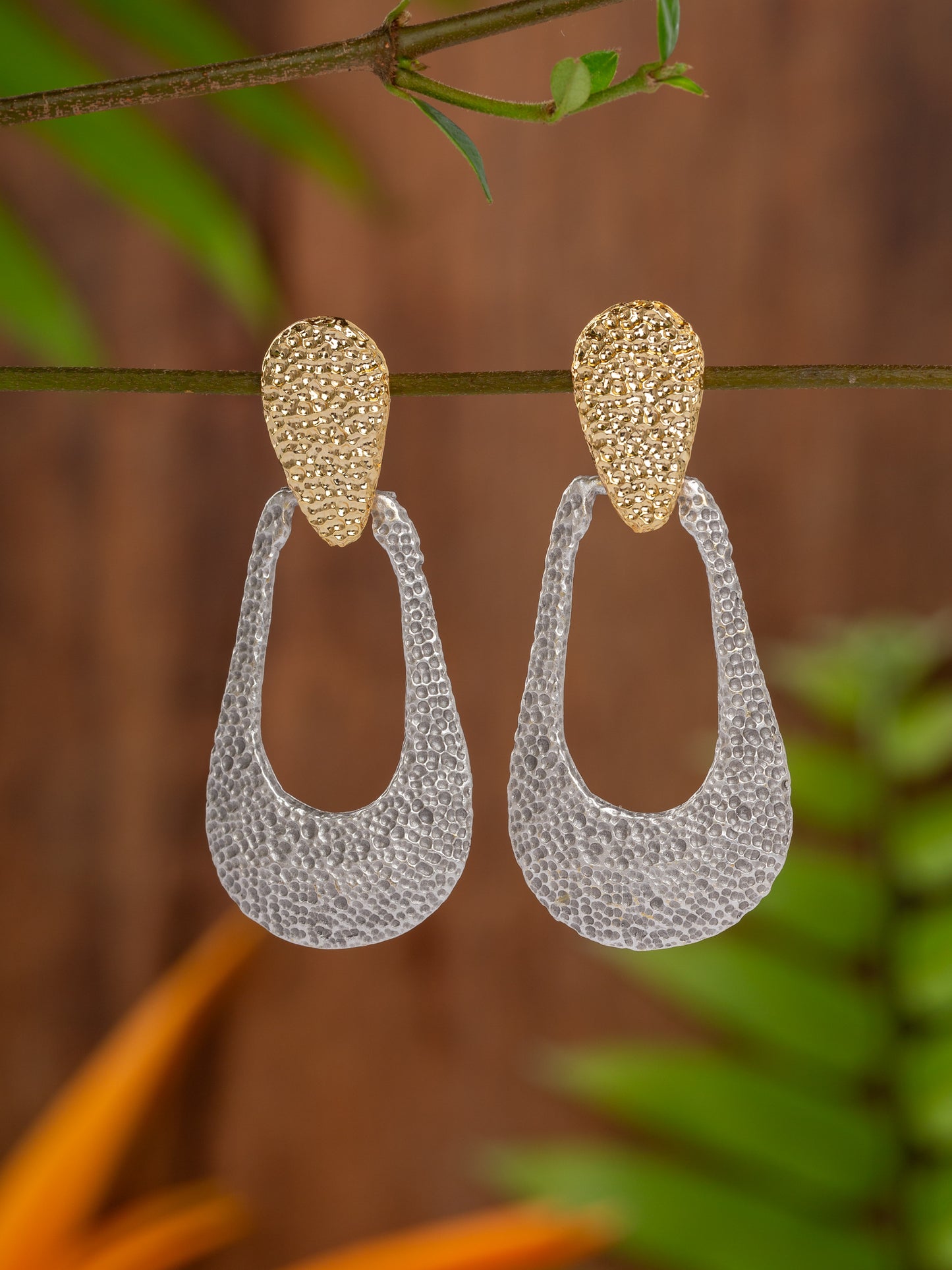 Gold and silver earrings in a raw beaten oval design
hanging on a branch with a blurred natural background