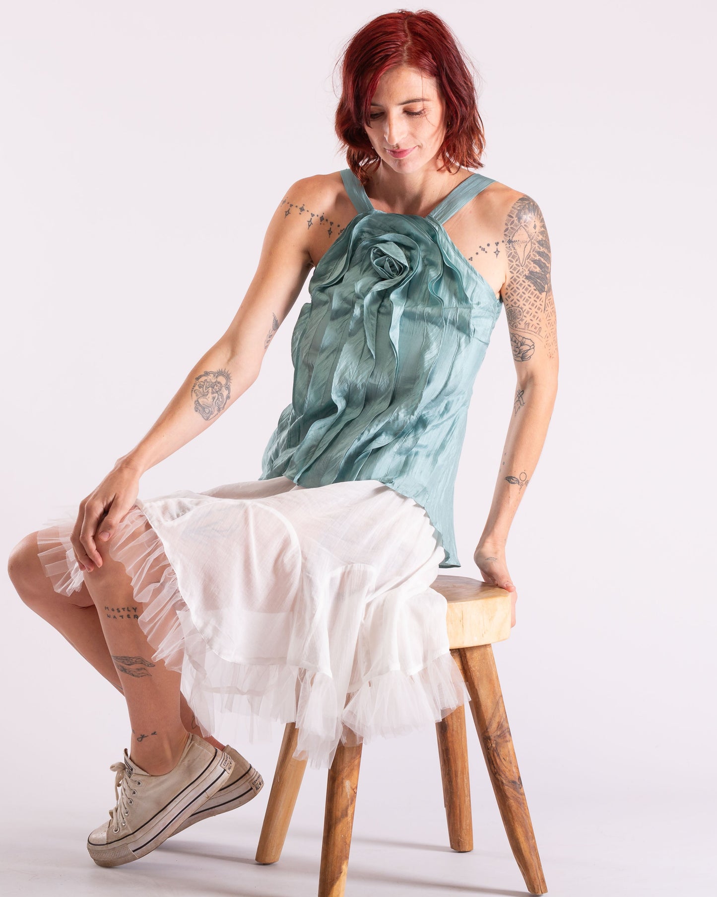 A woman with red hair sits on a wooden stool wearing a teal sleeveless top with a large rose detail and a white tiered skirt, against a plain light background.