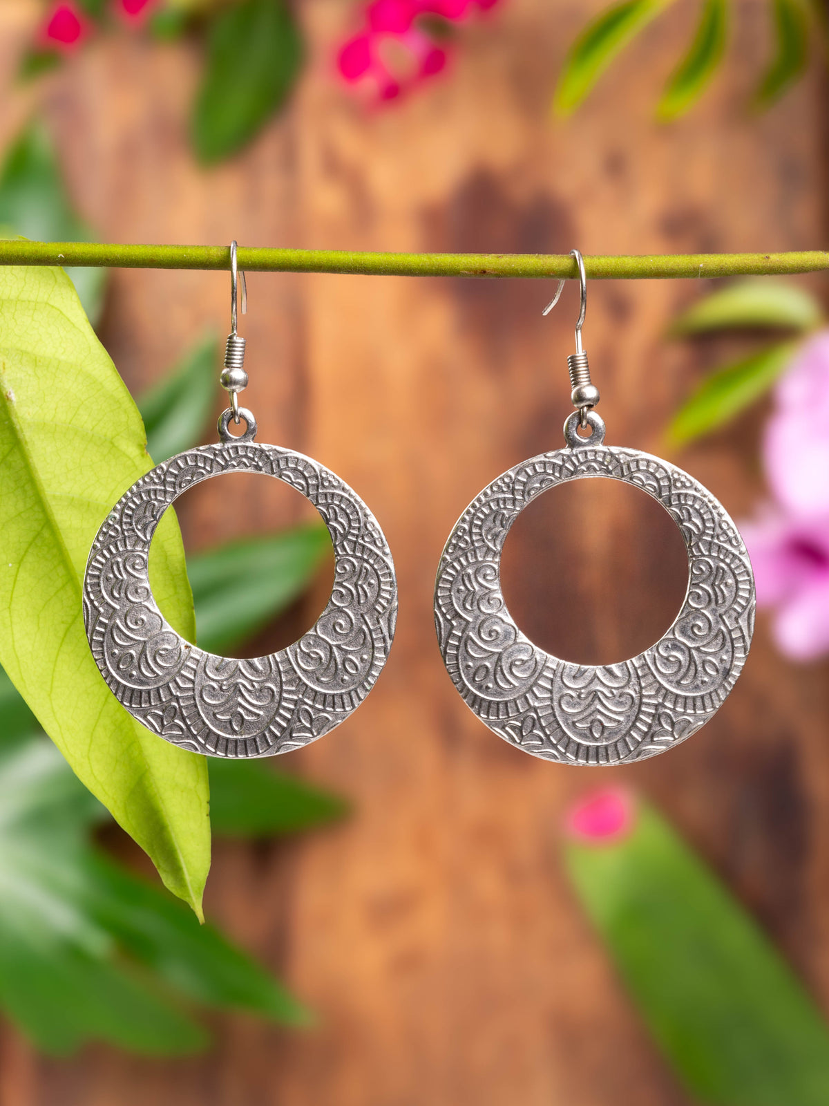 A pair of silver-colored Marley earrings with a geometric pattern, hanging from a wooden branch with green leaves in the background.