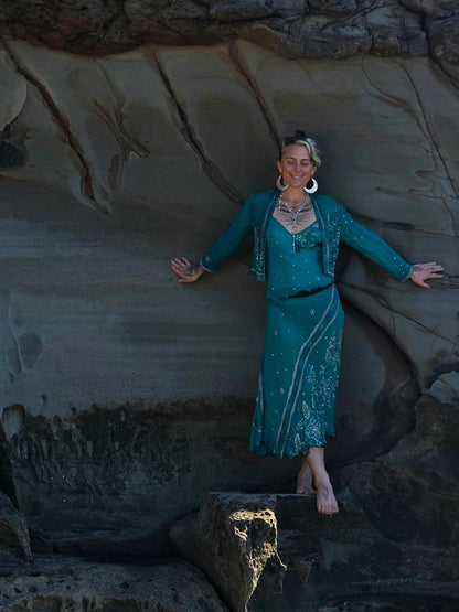A woman posing in a teal blue embroidered dress and matching jacket against a large rock formation.