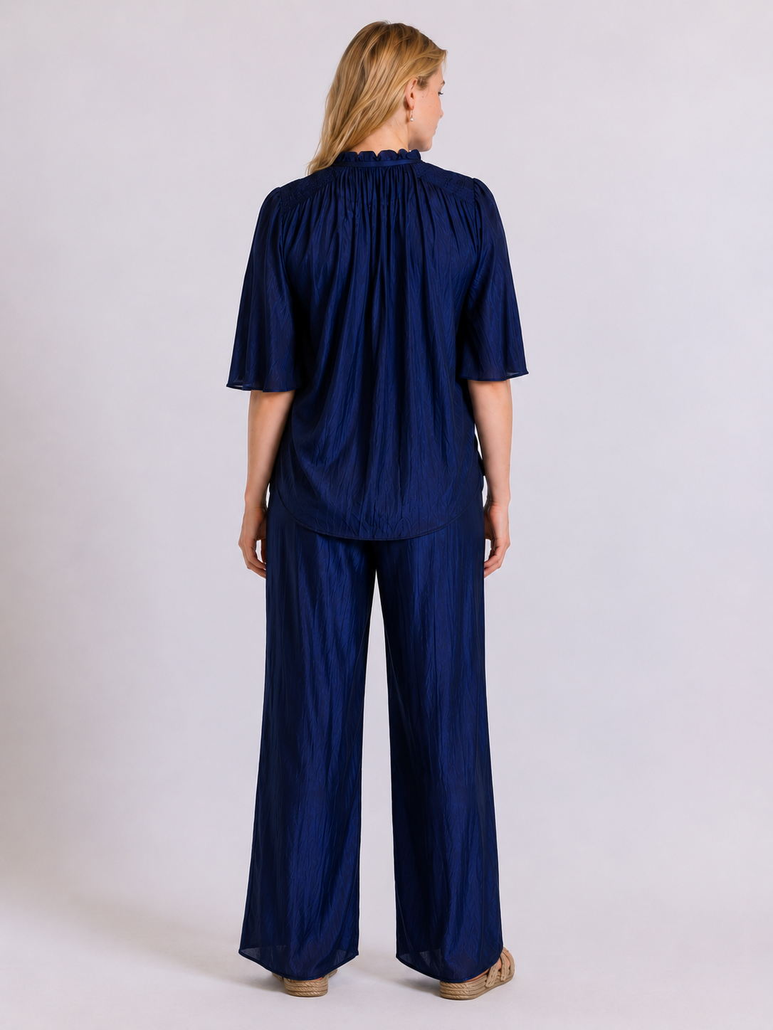A woman viewed from the back wearing a navy blue two-piece set consisting of a short-sleeve top with a gathered, pleated texture and matching wide-leg trousers, set against a plain light gray studio background.