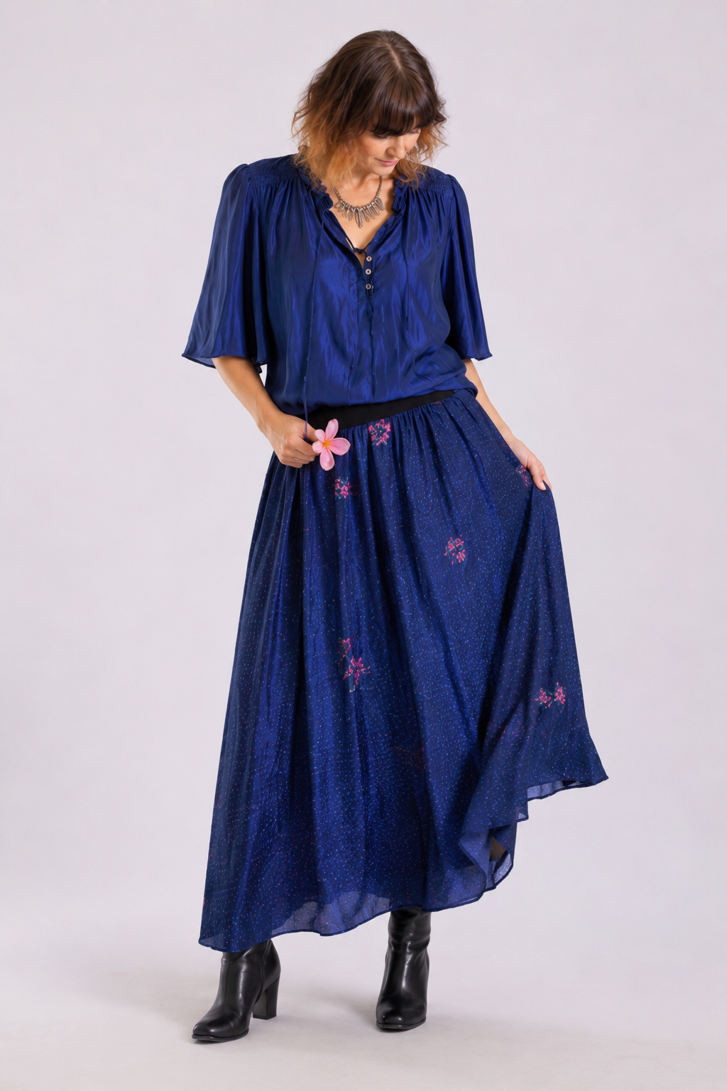 A woman modeling a two-piece navy blue set consisting of a short-sleeve button-up blouse and a matching high-waisted maxi skirt with pink floral embroidery, paired with black ankle boots.