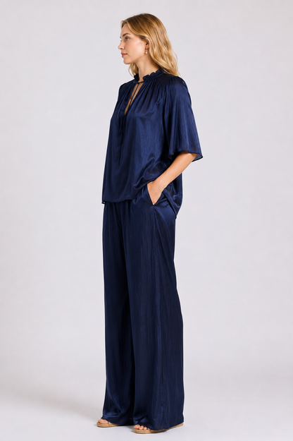 A woman modeling a navy blue silktwo-piece set consisting of a short-sleeve top with a split neckline and matching wide-leg pants, photographed against a light gray studio background.