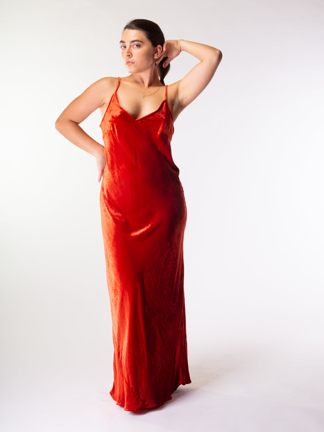 A woman modeling a floor-length red satin slip dress with thin spaghetti straps, posed against a seamless white studio background.