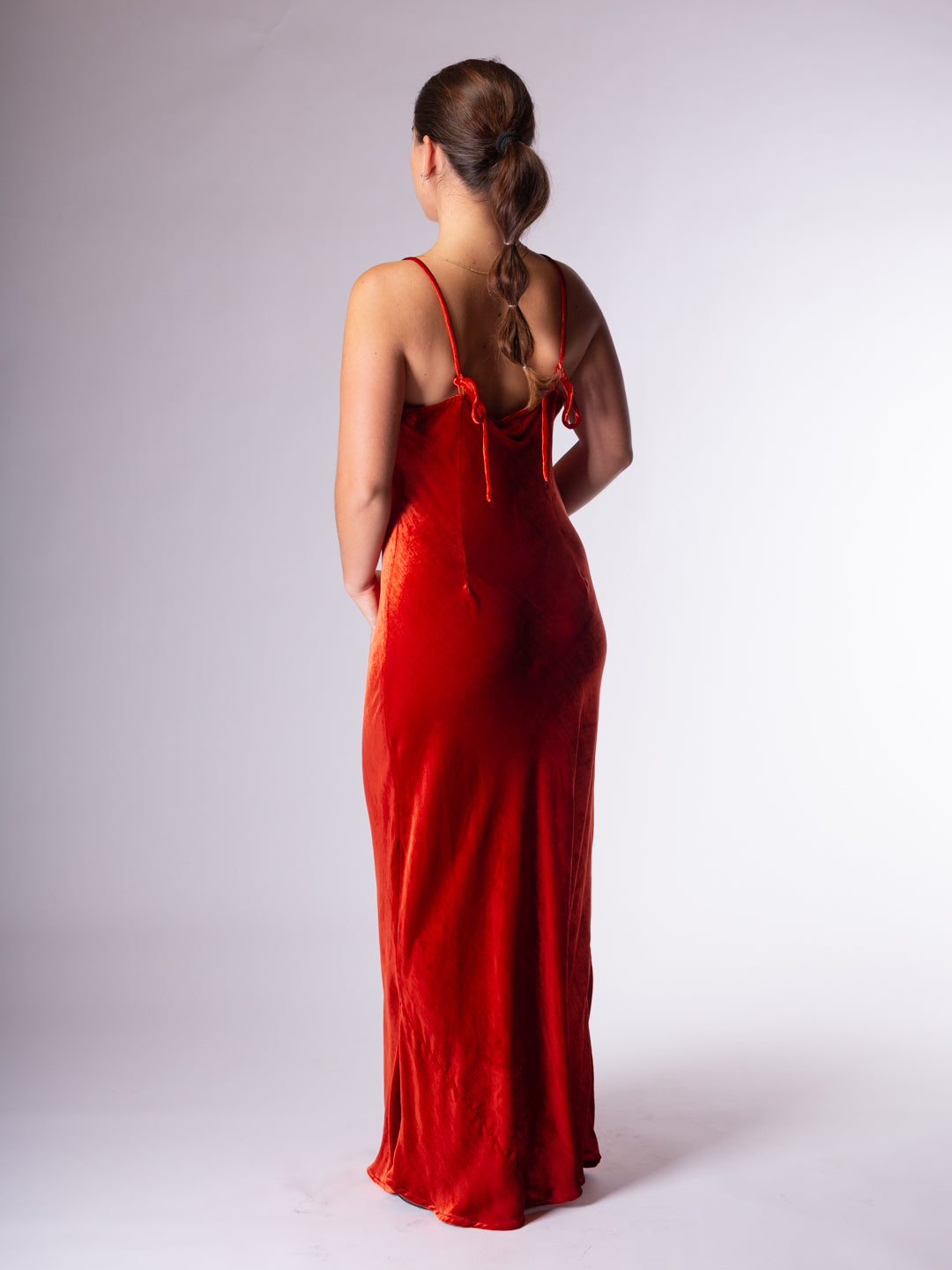 A woman viewed from behind wearing a long, sleeveless red dress with thin straps  photographed against a plain light gray studio background.