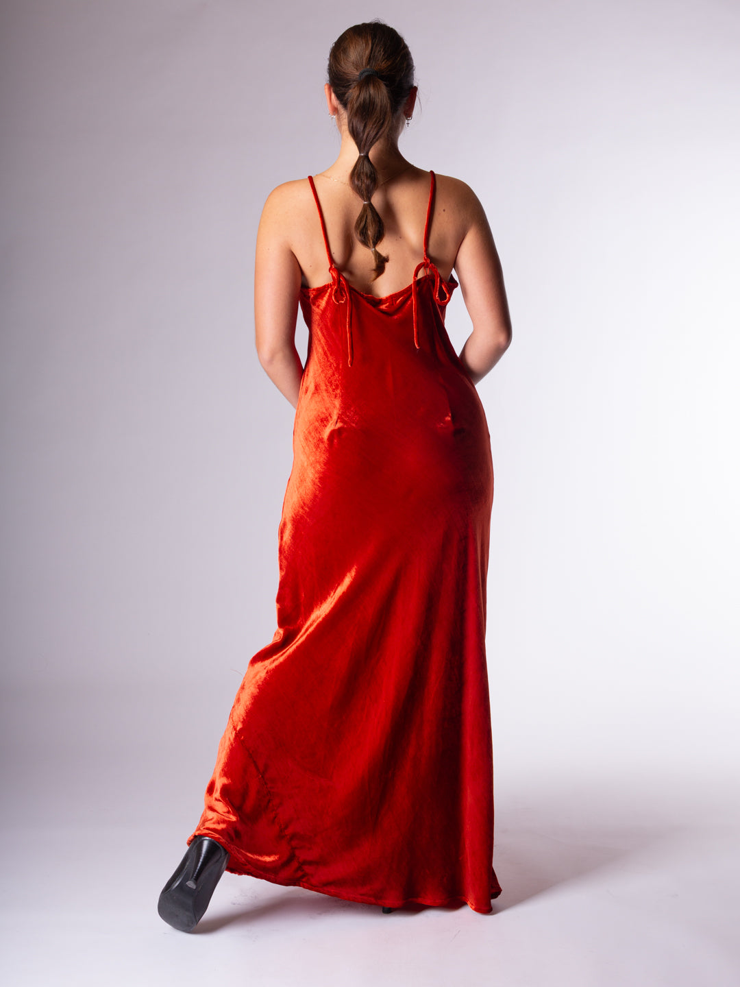 A woman viewed from behind wearing a long rust velvet slip dress with thin adjustable straps and a low open back, paired with black shoes, against a light gray studio background.
