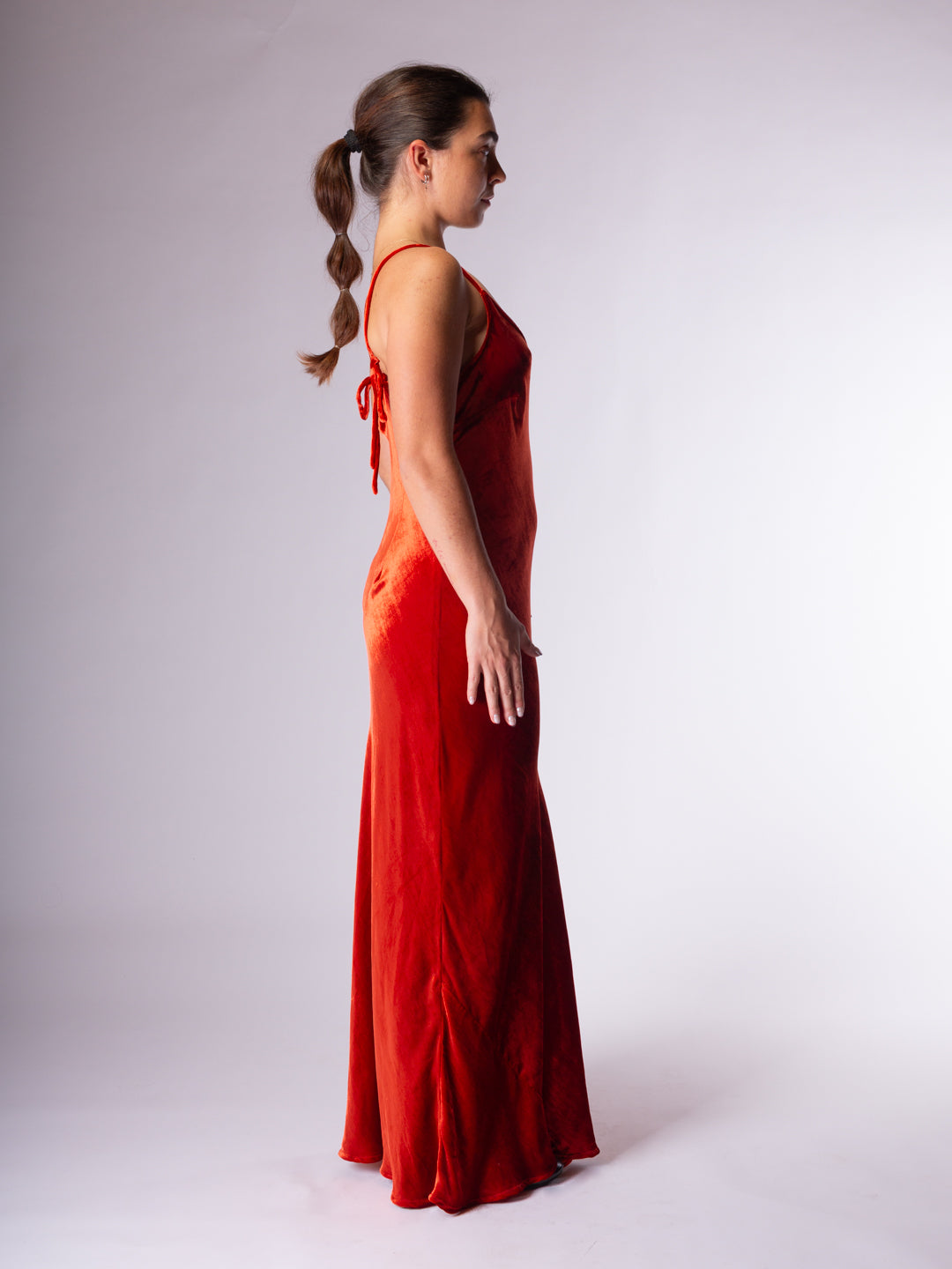 A woman in profile wearing a sleeveless, floor-length red dress with tie details at the back, photographed against a plain light gray studio background.