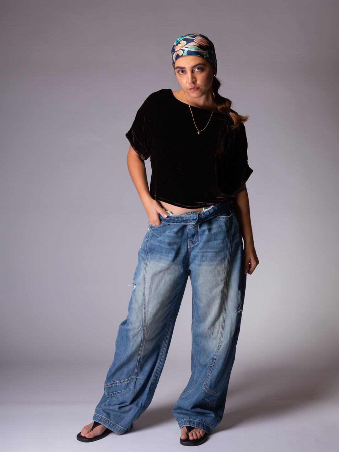Person wearing a dark chocolate t-shirt and blue jeans with a patterned headscarf on a gray background