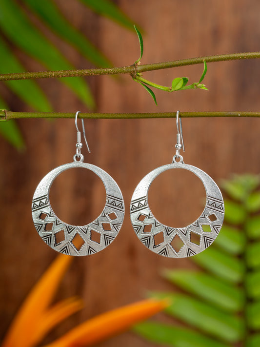 Silver hoop earrings with geometric patterns hanging on a branch with green leaves.