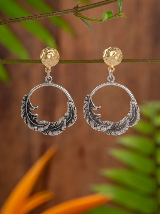 Silver feather-shaped earrings with gold accents on a branch with green leaves.