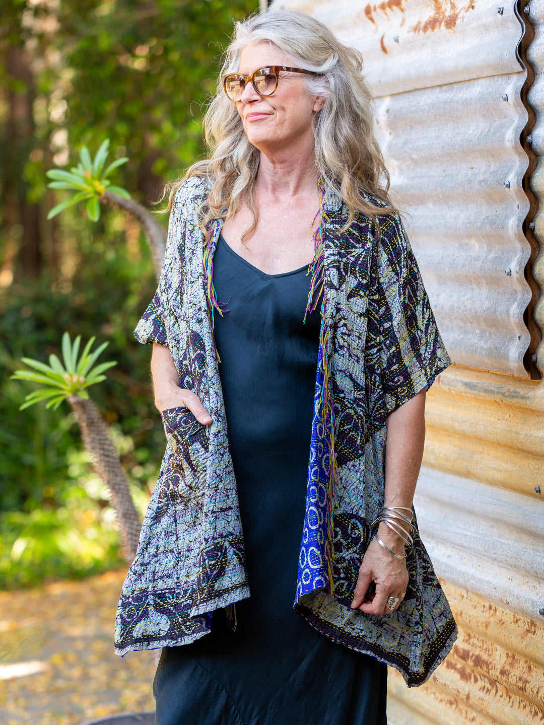 Woman wearing a patterned kimono over a dark dress, standing against a corrugated metal wall with greenery in the background.