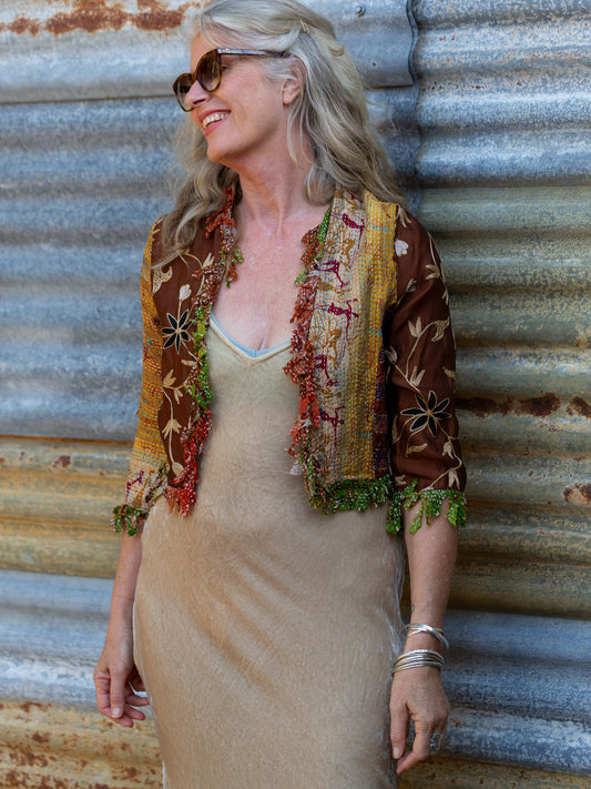 Woman in a patterned jacket and beige dress leaning against a corrugated metal wall.