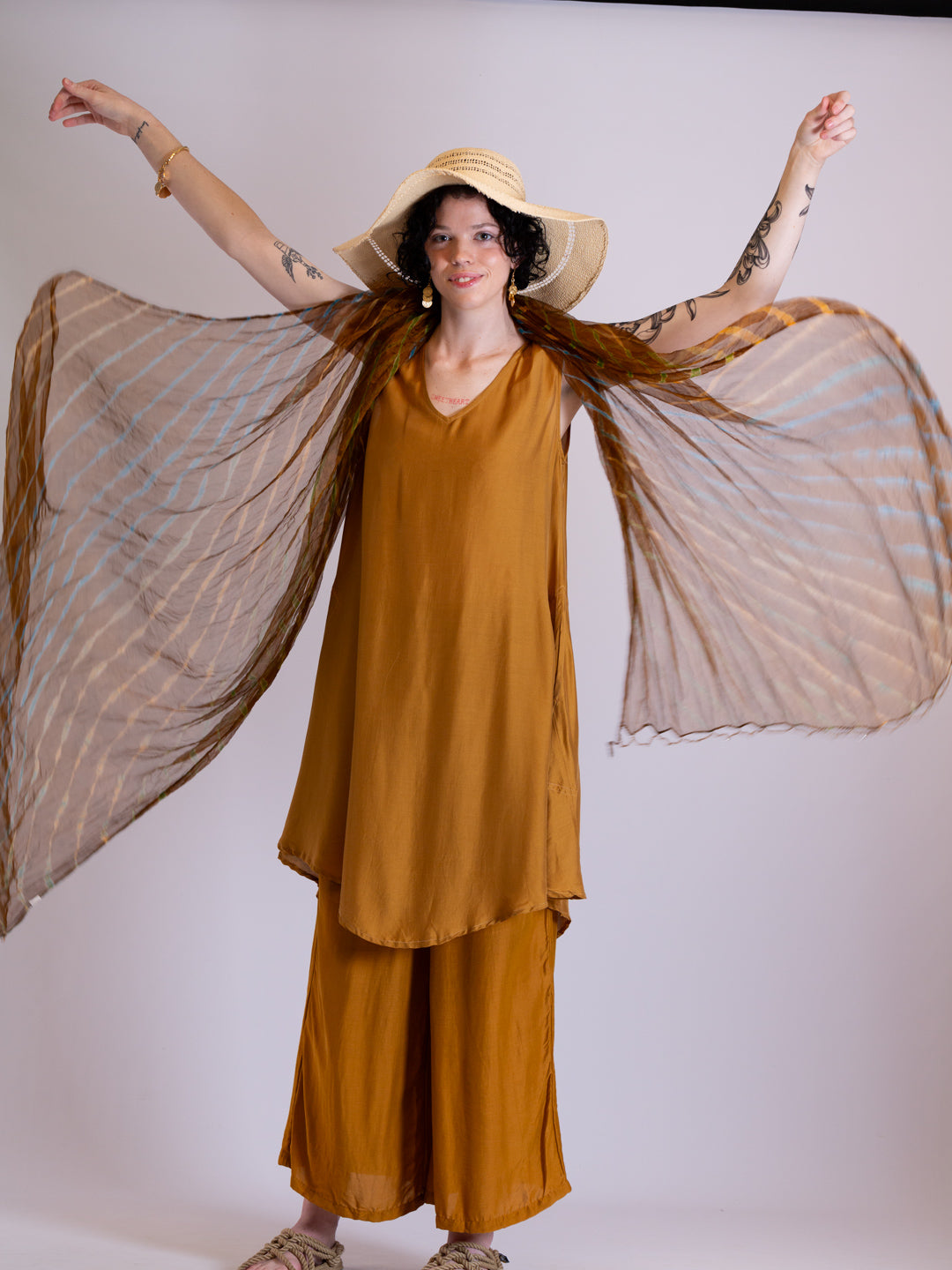 Woman in a mustard yellow dress and hat with a flowing scarf against a white background