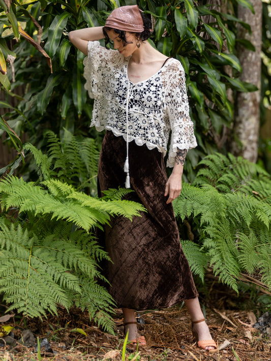 Woman in a white crochet lace top and brown skirt standing among green foliage