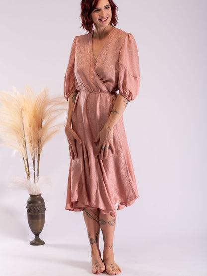 A woman modeling a dusty rose pink midi wrap dress with puff sleeves , standing next to a decorative vase with pampas grass against a light gray studio background.