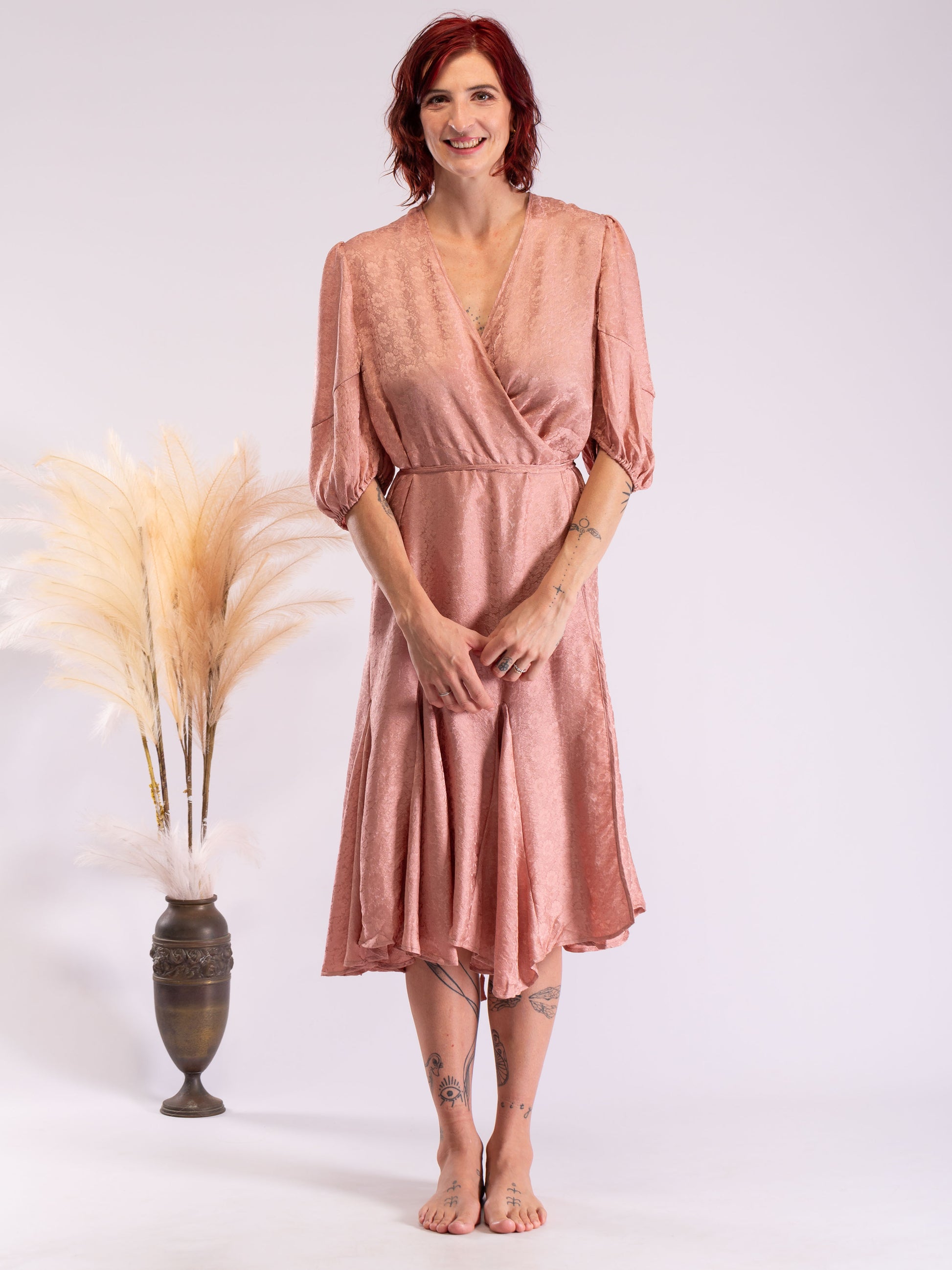 A woman with red hair stands barefoot in a studio wearing a dusty rose wrap midi dress with puff sleeves. To her left is a tall dark vase filled with pampas grass against a light gray background.