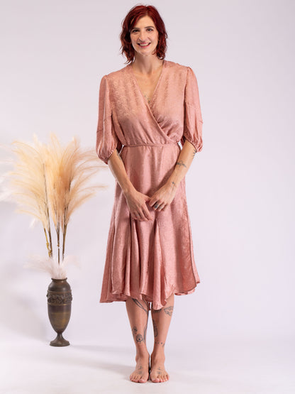 A woman with red hair stands barefoot in a studio wearing a dusty rose wrap midi dress with puff sleeves. To her left is a tall dark vase filled with pampas grass against a light gray background.