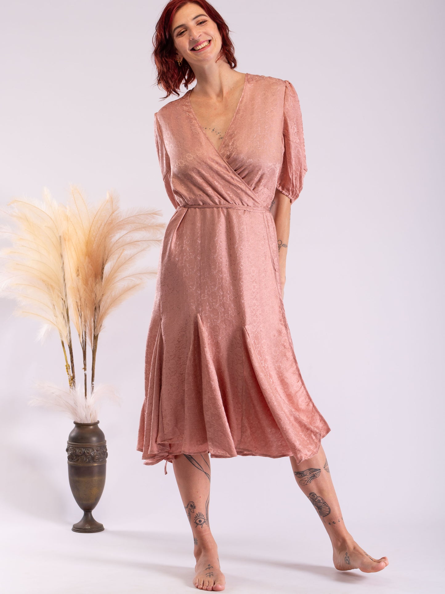 A woman modeling a dusty rose pink midi wrap dress with short puff sleeves and a V-neckline, standing barefoot next to a decorative vase with pampas grass against a light gray studio background.