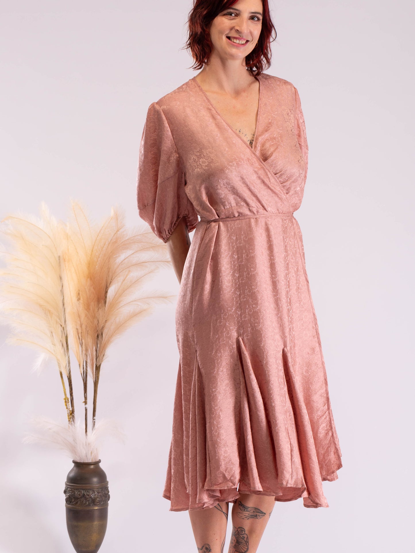 A woman wearing a dusty rose pink wrap midi dress with short puff sleeves and a textured shimmer finish, standing next to a decorative vase with pampas grass against a light gray studio background.