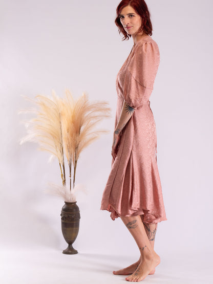 A woman wearing a dusty rose pink midi wrap dress with a textured pattern, standing barefoot next to a decorative vase with pampas grass against a light gray background...otomo lifestyle