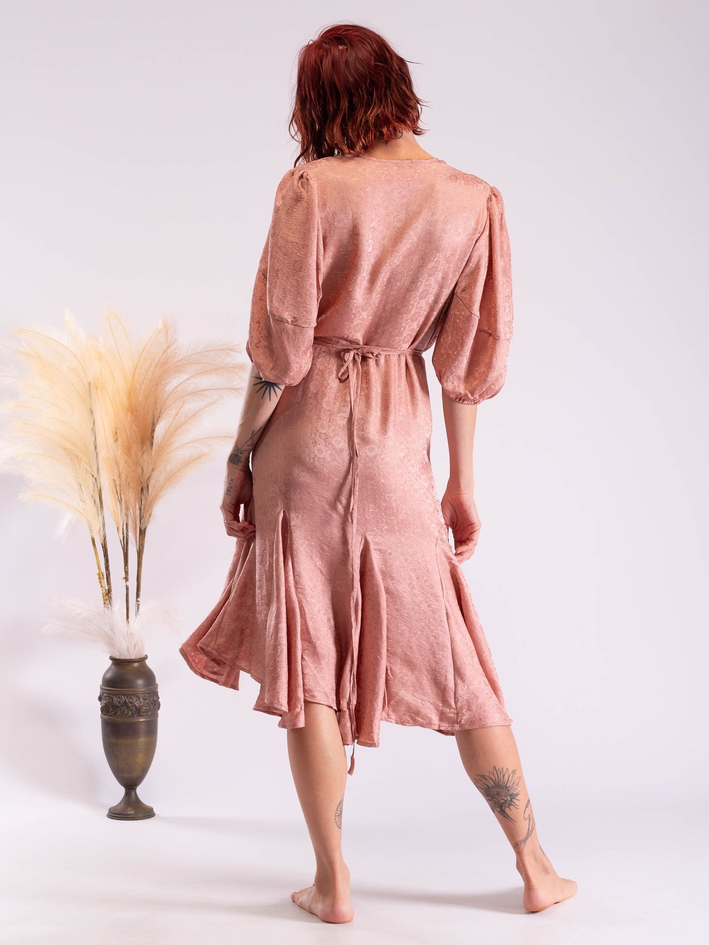 A woman with red hair is shown from behind wearing a dusty rose pink linen midi dress with puff sleeves and a tie waist. She is standing barefoot next to a decorative vase with dried pampas grass against a plain white studio background.