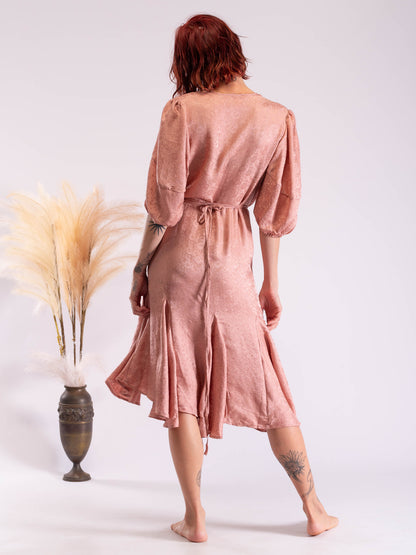 A woman with red hair is shown from behind wearing a dusty rose pink linen midi dress with puff sleeves and a tie waist. She is standing barefoot next to a decorative vase with dried pampas grass against a plain white studio background.