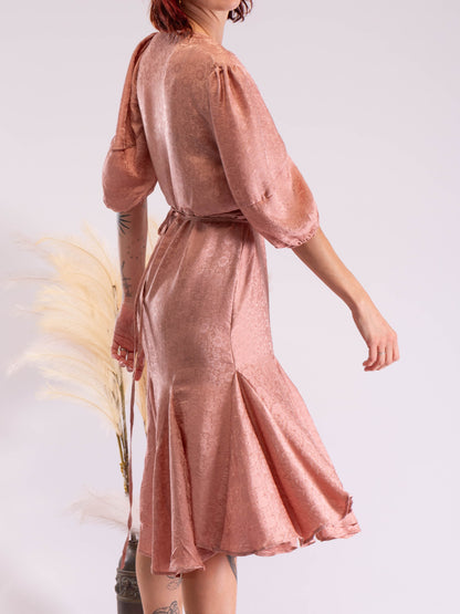 A woman wearing a shimmering dusty rose pink midi dress with puff sleeves and a tie waist, shown from a back three-quarter angle against a light studio background.