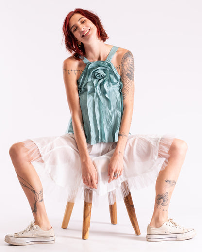 A model with red hair and tattoos sits on a wooden stool against a white studio background, wearing aduck egg blue silk halter top with a large rose detail, a white tulle skirt, and white canvas sneakers.