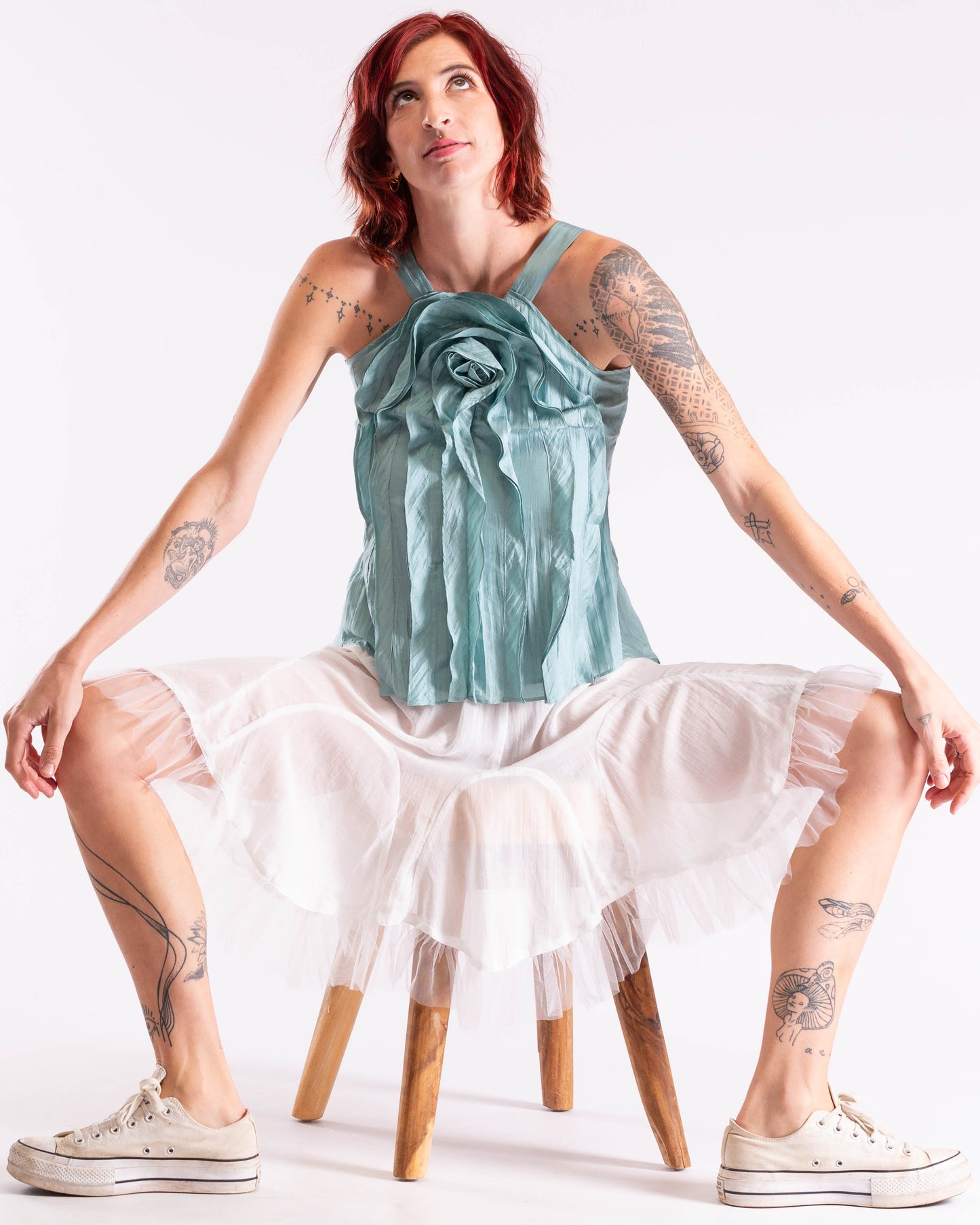 A woman with red hair and tattoos sits on a wooden stool wearing a light blue satin rose-shaped halter top and a white ruffled tulle skirt, paired with white canvas sneakers, against a plain white studio background.