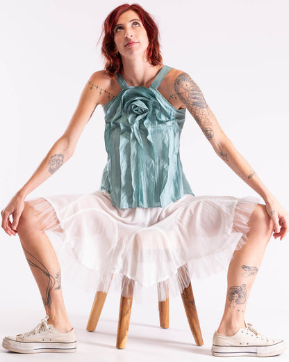 A woman with red hair and tattoos sits on a wooden stool wearing a light blue satin rose-shaped halter top and a white ruffled tulle skirt, paired with white canvas sneakers, against a plain white studio background.