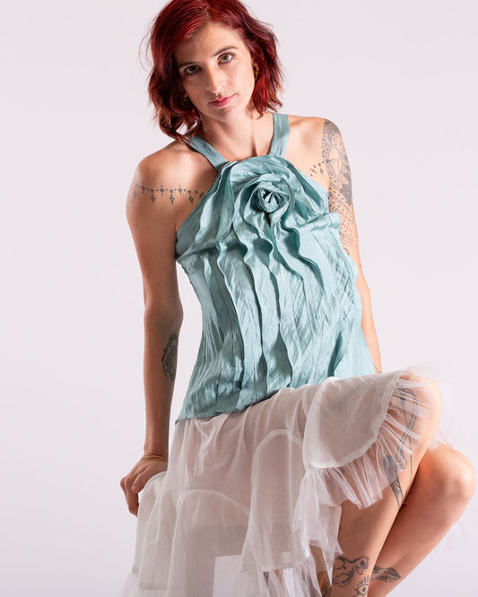 A woman with red hair and tattoos models a light blue halter top with a ruffled flower detail and a layered white tulle skirt against a plain white studio background.