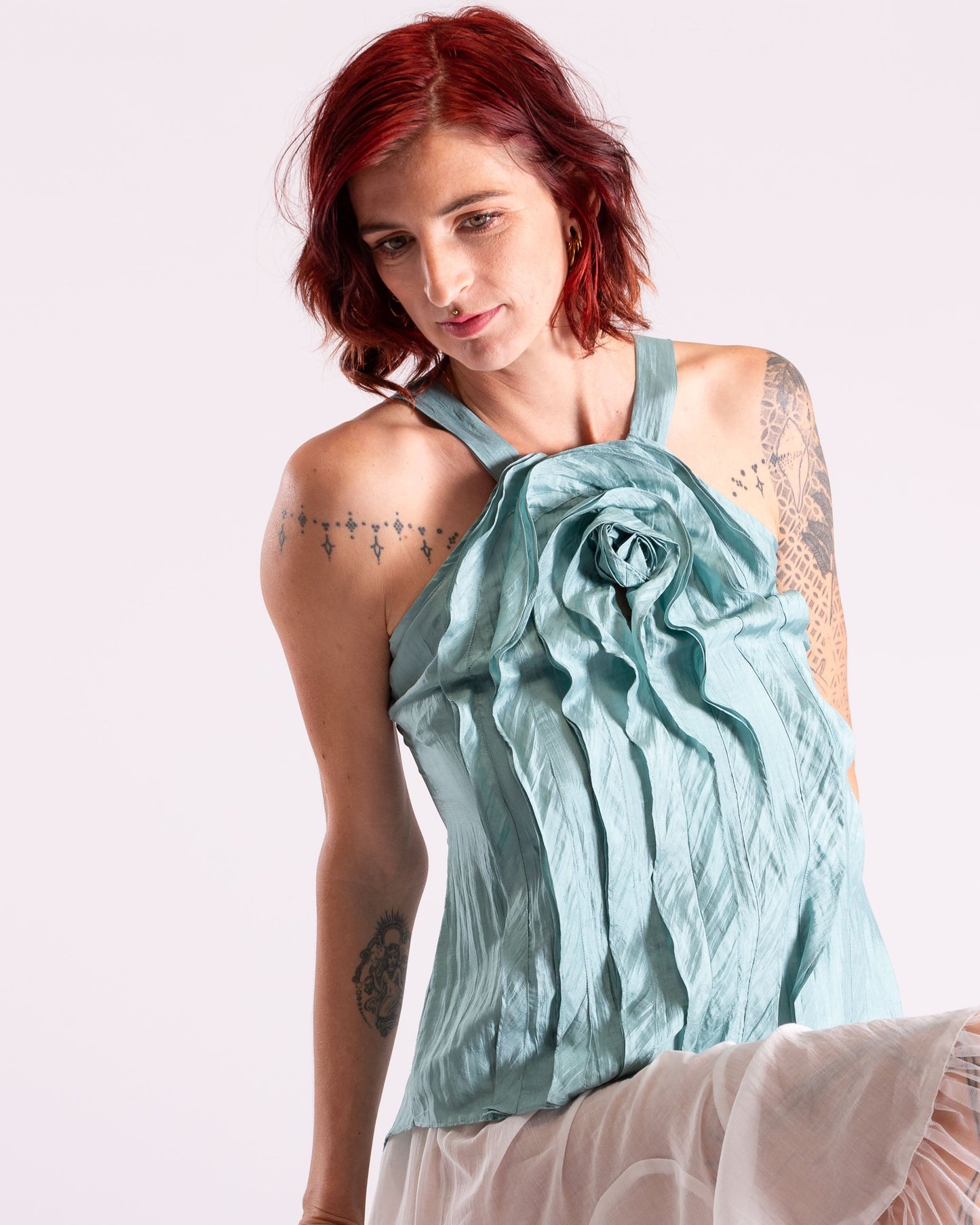A model with short red hair and tattoos wearing a light blue-green halter top with a large gathered rosette detail on the chest, paired with a sheer white skirt, against a plain white studio background.