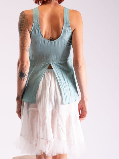 A woman shown from the back wearing a sleeveless dusty blue-green top with a deep open back and a layered white tulle skirt, against a plain light background.