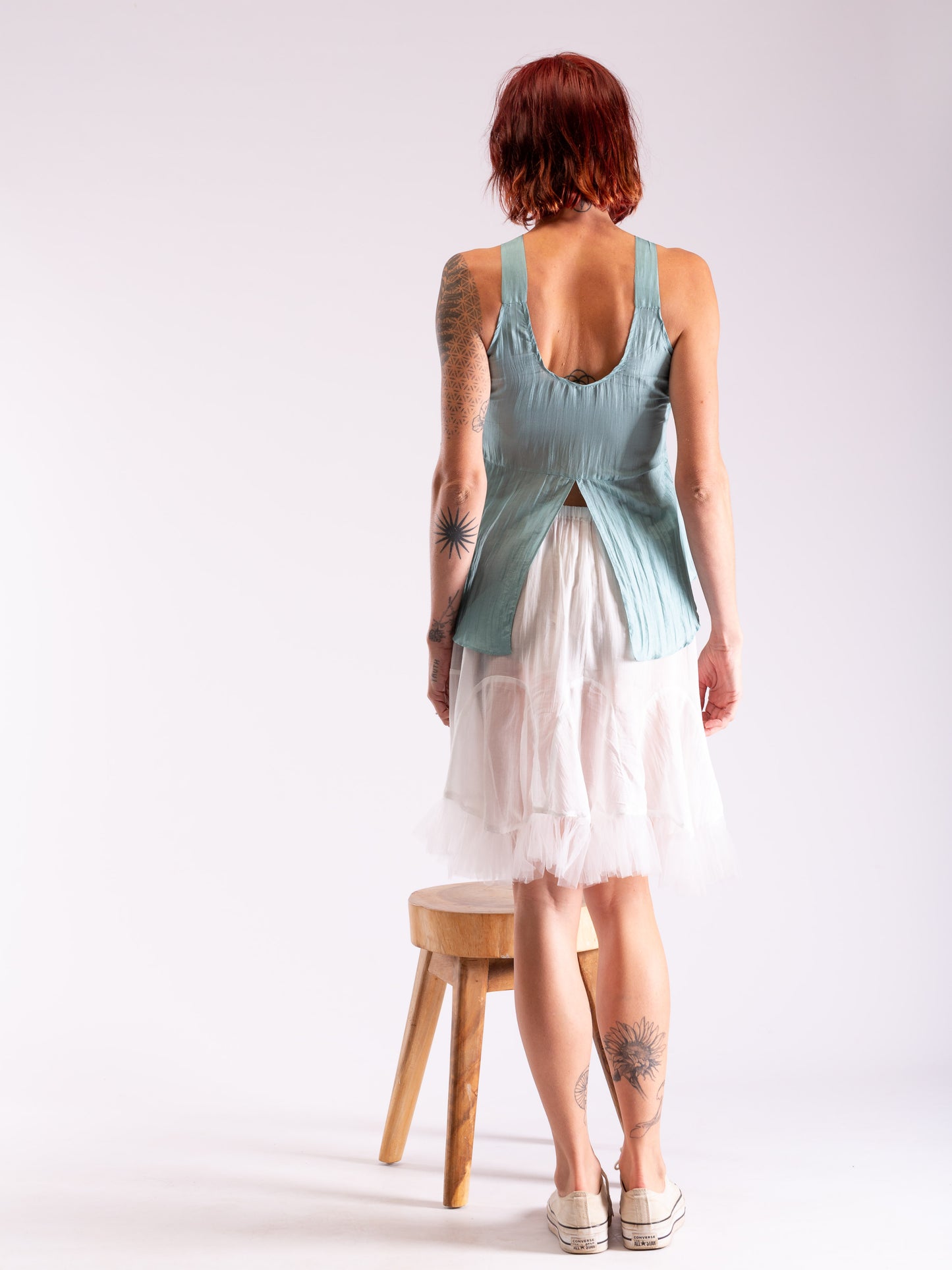 A woman with short red hair is shown from behind wearing a sleeveless sage green top and a white tiered tulle skirt, standing next to a small wooden stool against a plain white studio background.