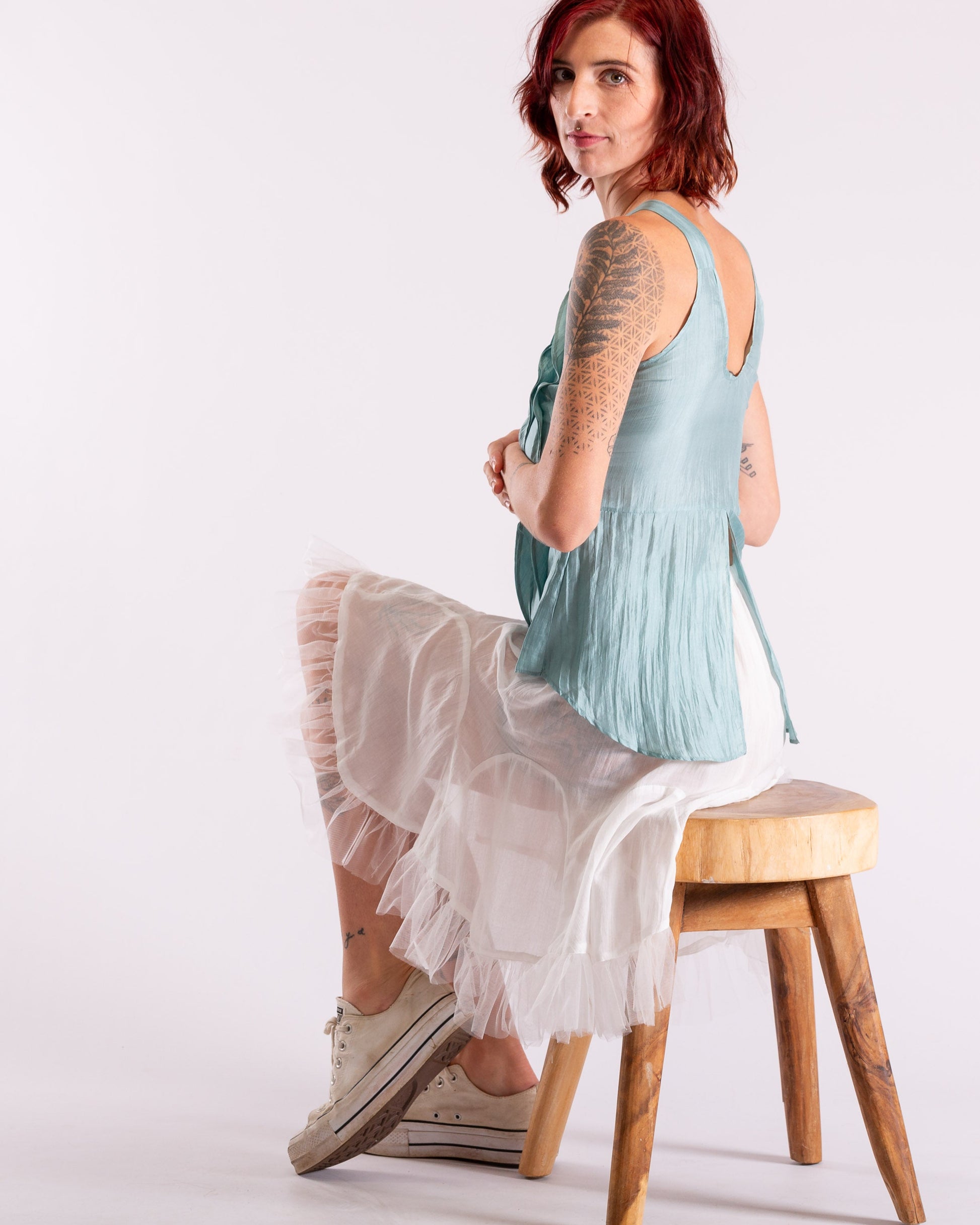 A woman with red hair sits on a wooden stool, wearing a light blue sleeveless top and a long white tulle skirt against a plain white studio background, otomo paris top