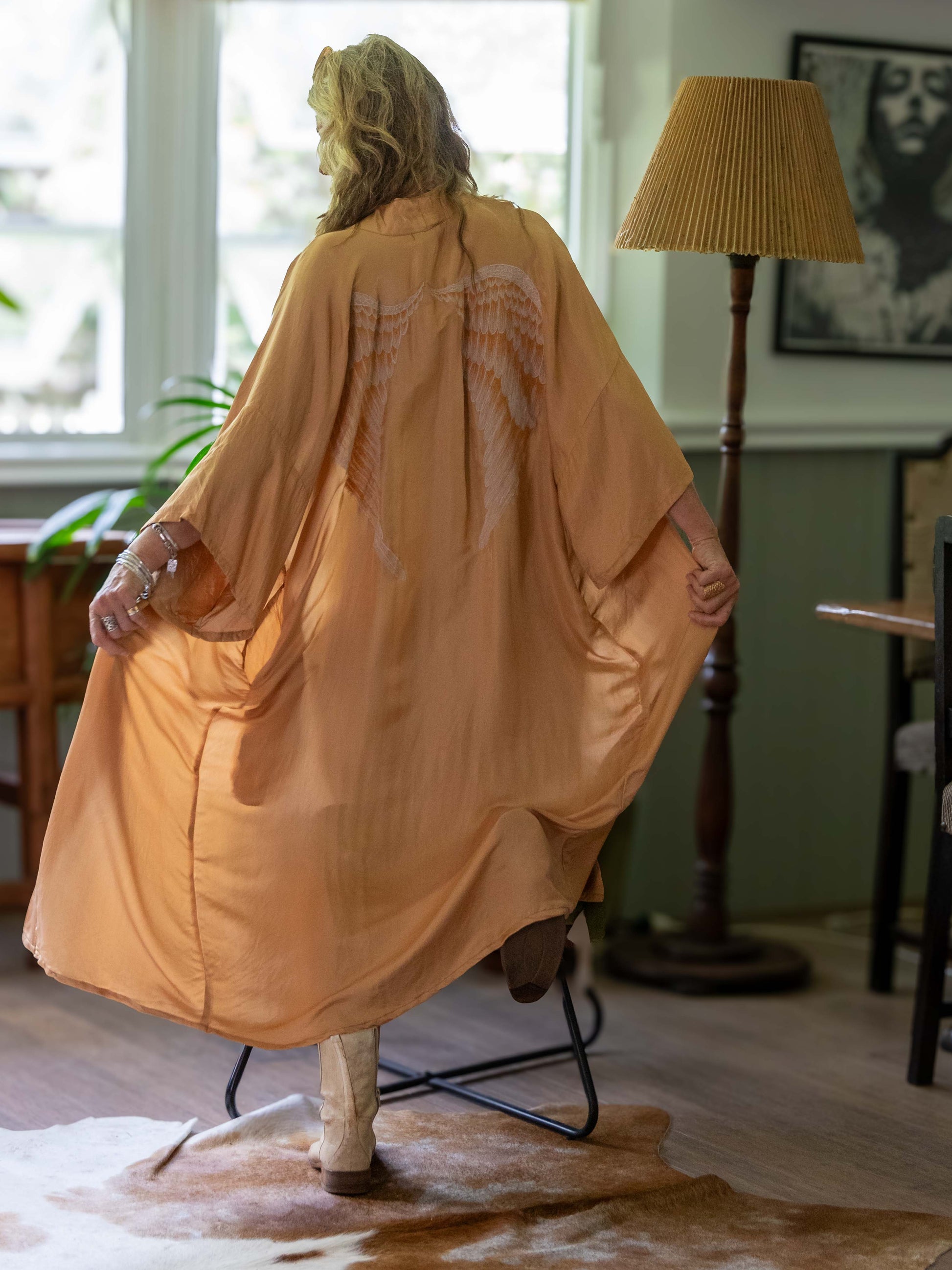 Person wearing a long, champagne kimono in a room with a lamp and window.