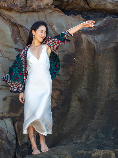 A woman stands barefoot on a rocky cliffside wearing a white satin slip dress, a colorful patterned kimono, and earrings, with her arm extended to the side.