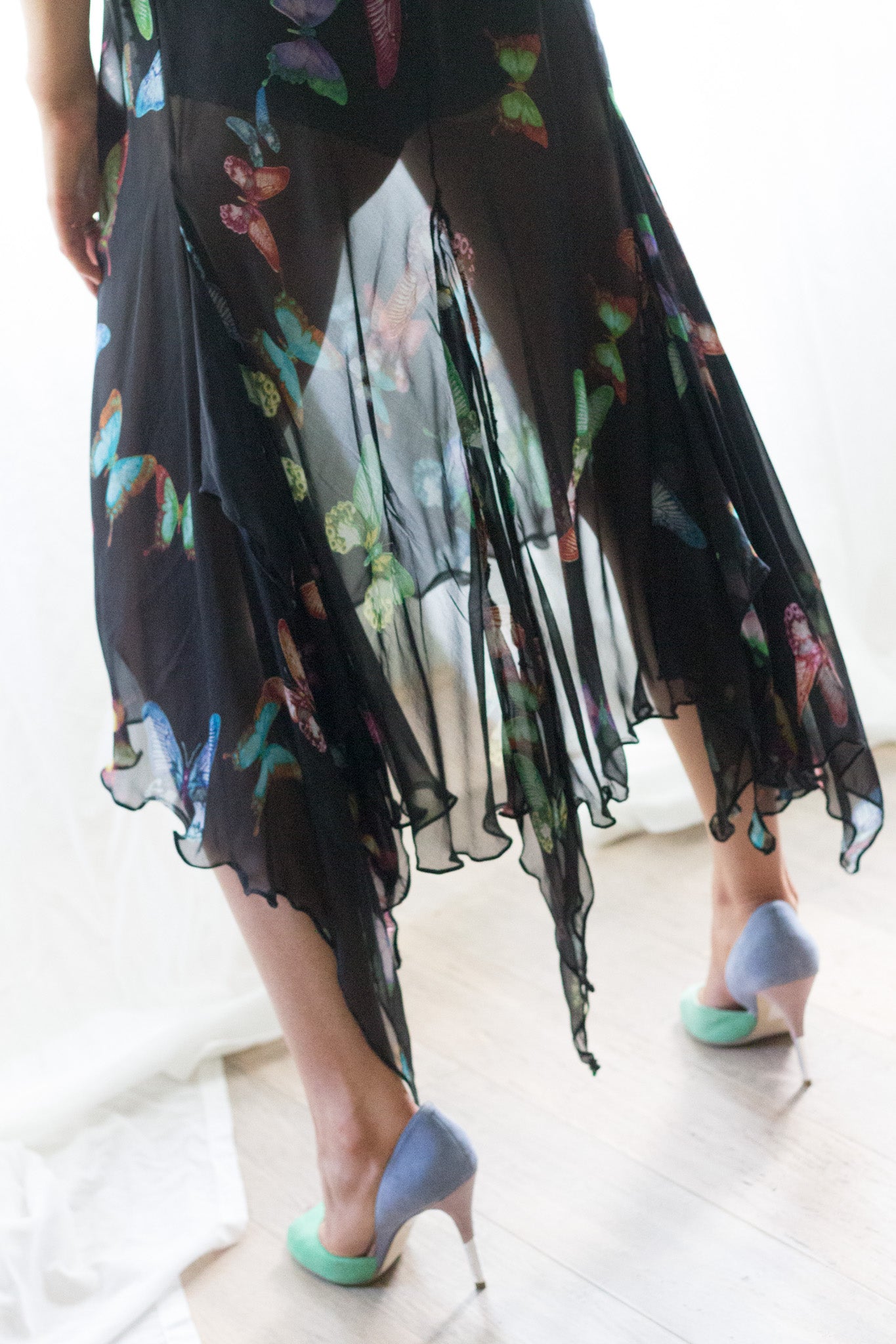 A person wearing a sheer black midi dresswith a colorful butterfly print, paired with light blue high heels, standing on a light wood floor.