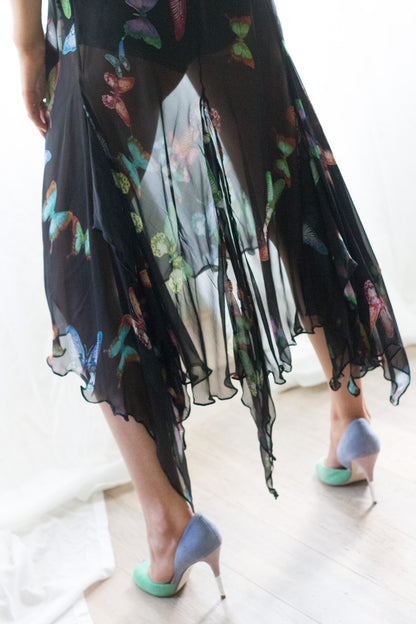 A person wearing a sheer black midi dresswith a colorful butterfly print, paired with light blue high heels, standing on a light wood floor.