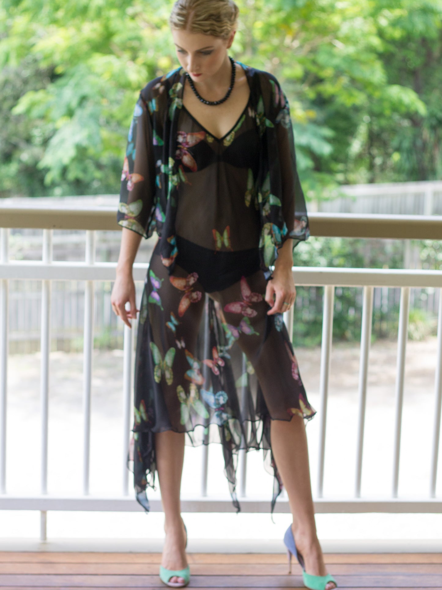 A woman stands on a balcony wearing a sheer silk georgette black midi dress with a myriad of butterflies layered over a black camisole and underwear set, paired with mint green high heels.
