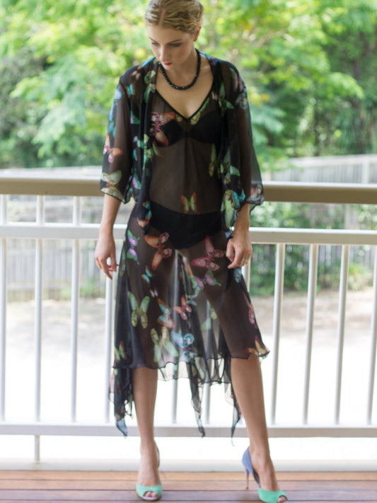 A woman stands on a balcony wearing a sheer silk georgette black midi dress with a myriad of butterflies layered over a black camisole and underwear set, paired with mint green high heels.