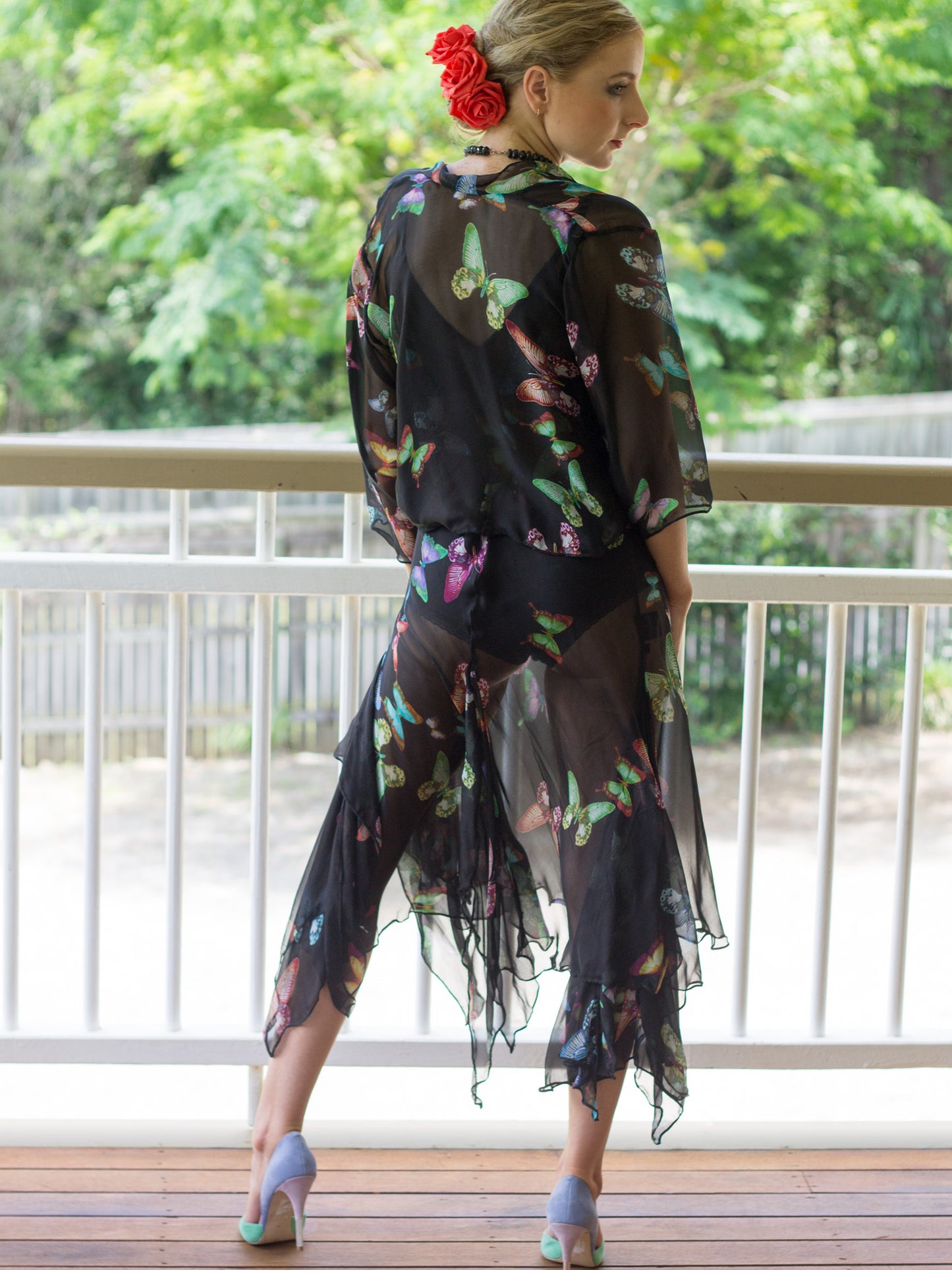 A woman stands on a balcony wearing a sheer black butterfly-print silk jacket and slip dress. The fabric has a colorful butterfly pattern with green, pink, and blue tones.