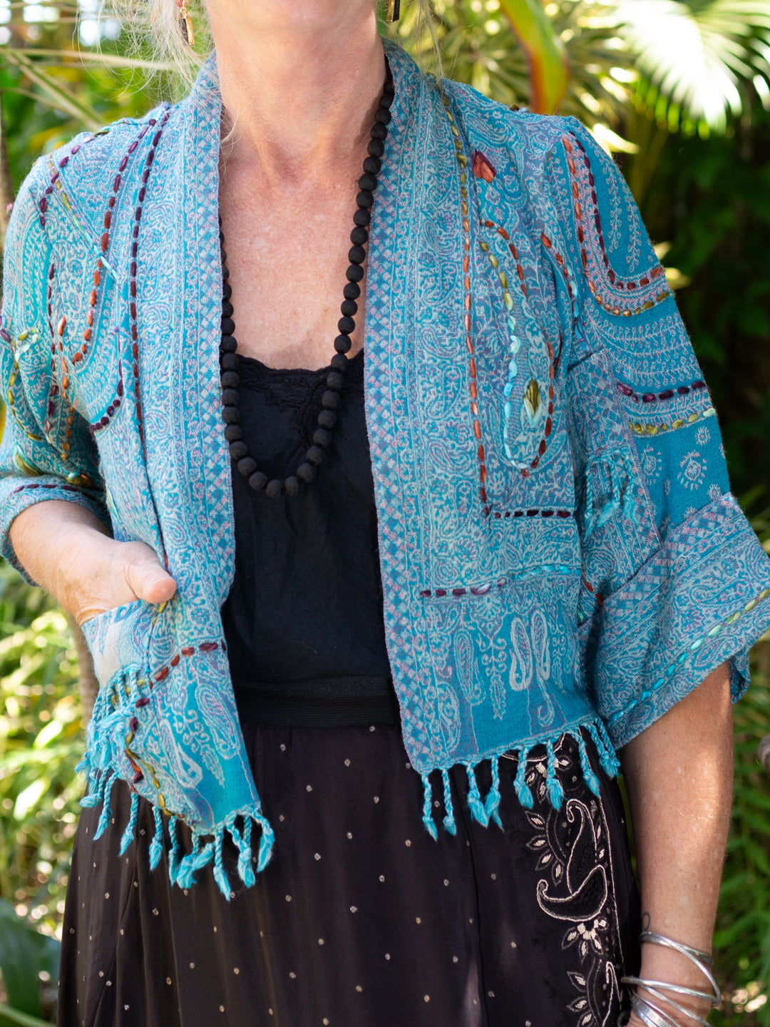 Person wearing a blue embroidered jacket with a black necklace against a green leafy background