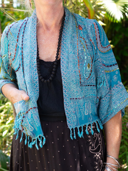 Person wearing a blue embroidered jacket with a black necklace against a green leafy background