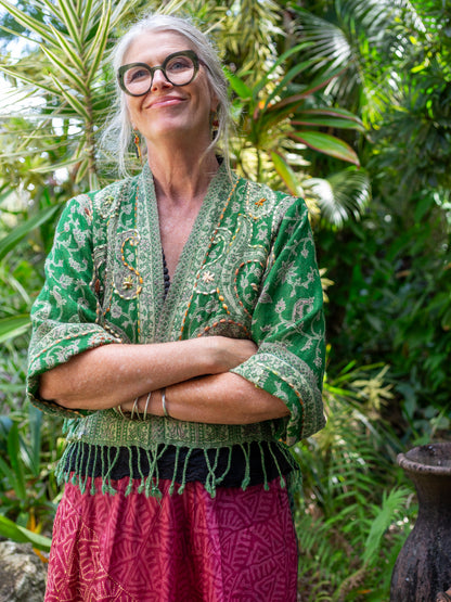 Person wearing a green patterned top and pink skirt standing in a lush, green outdoor setting.