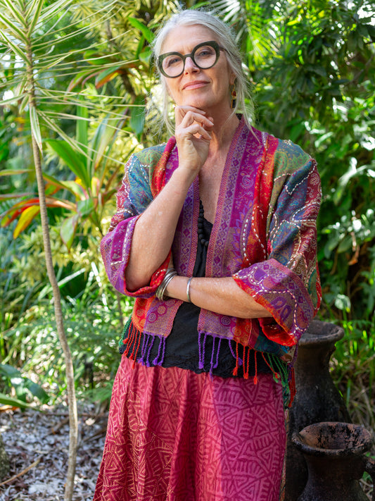 Woman in a colorful jacket and pink dress standing in a lush green forest.