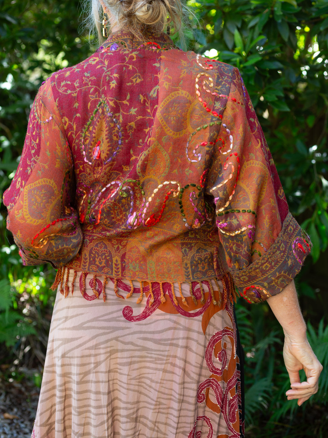 Person wearing a patterned outfit with a red and orange top and beige skirt against a green leafy background