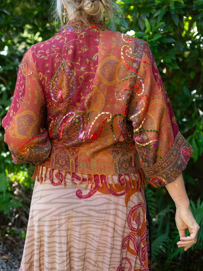 Person wearing a patterned outfit with a red and orange top and beige skirt against a green leafy background