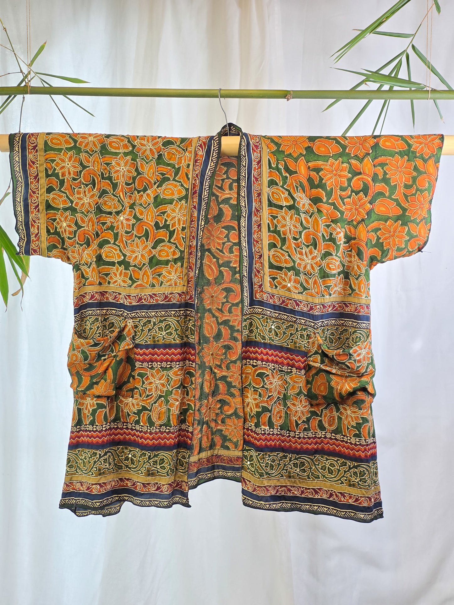 A short-sleeved open-front kimono-style top with a bold orange and green floral print, hanging on a bamboo rod against a white curtain backdrop with decorative bamboo leaves.